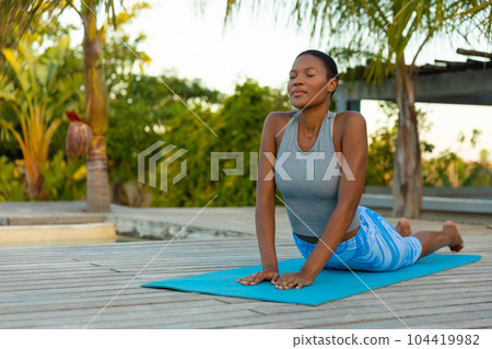 Happy african american woman practicing yoga stretching with eyes closed on beach deck, copy space Happy african american woman practicing yoga stretching with eyes closed on beach deck, copy space 104419982