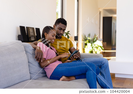 Happy african american couple smiling, embracing and using smartphone on couch at home Happy african american couple smiling, embracing and using smartphone on couch at home 104420004