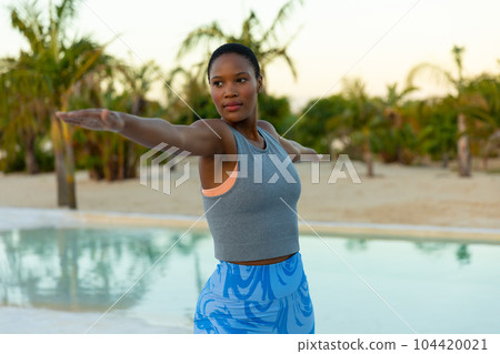 Happy african american woman practicing yoga standing and stretching by pool on beach at sundown 104420021