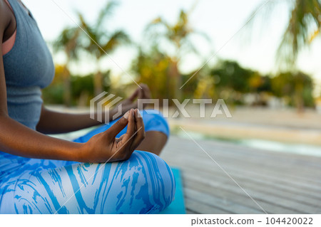 Midsection of african american woman practicing yoga meditation sitting on sunny beach 104420022