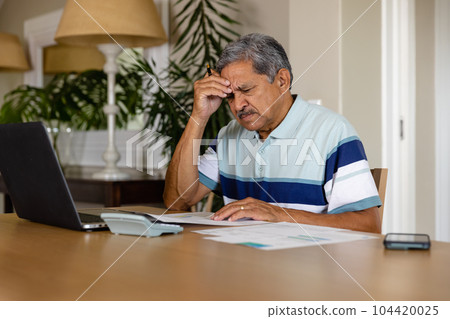 Senior biracial man doing paperwork and using laptop in dining room Senior biracial man doing paperwork and using laptop in dining room 104420025