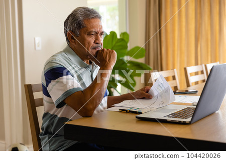 Senior biracial man doing paperwork and using laptop in dining room Senior biracial man doing paperwork and using laptop in dining room 104420026