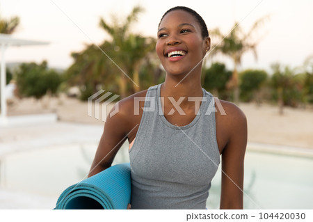 Happy african american woman smiling and holding yoga mat on sunny beach Happy african american woman smiling and holding yoga mat on sunny beach 104420030