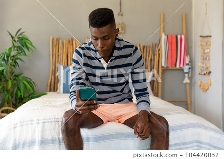 African american man sitting on bed using smartphone at holiday beach house African american man sitting on bed using smartphone at holiday beach house 104420032