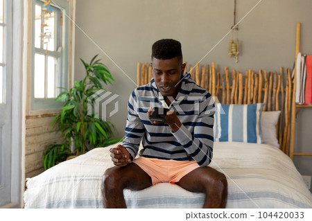 Happy african american man sitting on bed talking on smartphone at sunny holiday beach house Happy african american man sitting on bed talking on smartphone at sunny holiday beach house 104420033