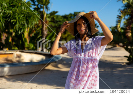 Portrait of happy biracial woman in white beach dress and sunhat smiling on sunny beach 104420034