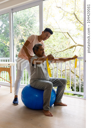 Diverse male physiotherapist advising and senior male patient using exercise band 104420081