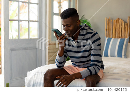 Happy african american man sitting on bed talking on smartphone at sunny holiday beach house Happy african american man sitting on bed talking on smartphone at sunny holiday beach house 104420153