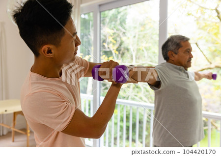 Diverse male physiotherapist advising and determined senior male patient using dumbbells 104420170