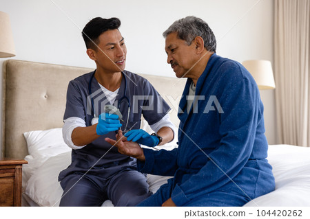 Diverse male doctor testing blood of senior male patient at home 104420262