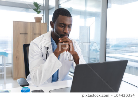 African american male doctor wearing lab coat and stethoscope, using laptop at hospital African american male doctor wearing lab coat and stethoscope, using laptop at hospital 104420354