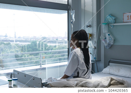 African american female patient sitting on bed and looking through window in hospital room African american female patient sitting on bed and looking through window in hospital room 104420365