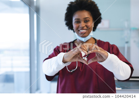 Portrait of happy african american female doctor wearing scrubs, forming hands in heart shape Portrait of happy african american female doctor wearing scrubs, forming hands in heart shape 104420414