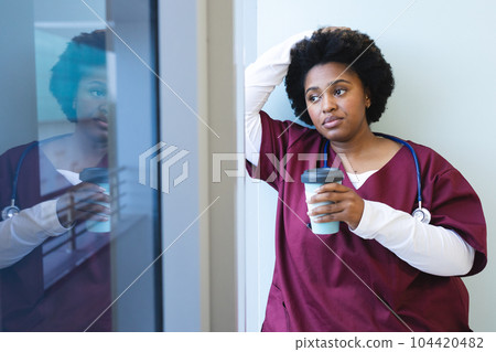 Thoughtful african american female doctor wearing scrubs, holding coffee and looking through window Thoughtful african american female doctor wearing scrubs, holding coffee and looking through window 104420482