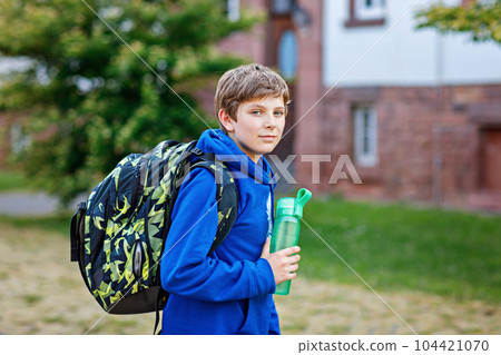 Happy preteen kid boy with backpack or satchel and water bottle. Schoolkid in on the way to elementary or middle school on warm sunny summer day. Healthy child outdoors on the street in the city 104421070