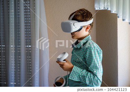 Young kid boy play VR games in virtual reality helmets at home. Exceted preteen child hold controllers and look away. Teenager in VR helmets playing video games. Young kid boy play VR games in virtual reality helmets at home. Exceted preteen child hold controllers and look away. Teenager in VR helmets playing video games. 104421123
