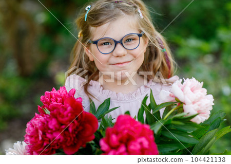 Cute adorable little preschool girl with huge bouquet of blossoming red and pink peony flowers. Portrait of smiling preschool child in domestic garden on warm spring or summer day. Summertime. 104421138