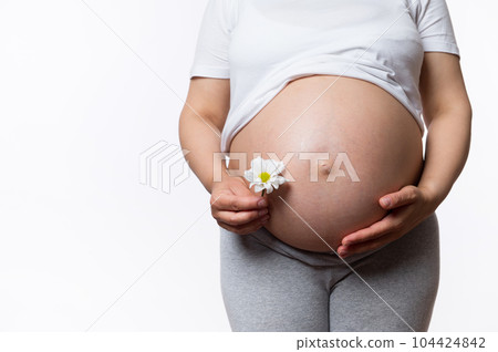 Pregnancy, maternity, childbearing and child birth concept. Close-up abdomen of a pregnant woman, expectant mother holding daisy flower near her belly. Obstetrics and gynecology. Fertility and health Pregnancy, maternity, childbearing and child birth concept. Close-up abdomen of a pregnant woman, expectant mother holding daisy flower near her belly. Obstetrics and gynecology. Fertility and health 104424842