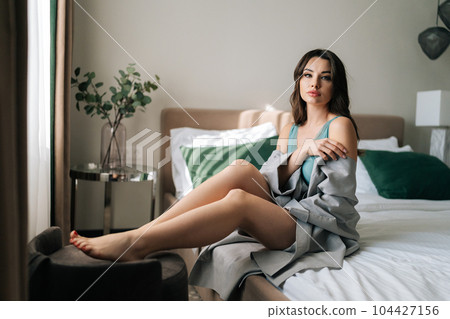 Portrait of gorgeous brunette female in sexy robe with perfect makeup and hairstyle sitting on bed looking at camera. Pretty young woman in gorgeous underwear posing in luxury bedroom in hotel. 104427156