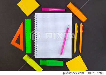 Back to school. On black table lies notebook checkered with pencil for text with empty space. All around are green, yellow, pink, orange stationery. Rulers, markers, felt tip pens, pens, adhesive note Back to school. On black table lies notebook checkered with pencil for text with empty space. All around are green, yellow, pink, orange stationery. Rulers, markers, felt tip pens, pens, adhesive note 104427780