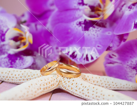 Golden wedding rings on white starfish behind purple orchids on pink wooden board. Celebrations, ceremonies, honeymoon 104427887