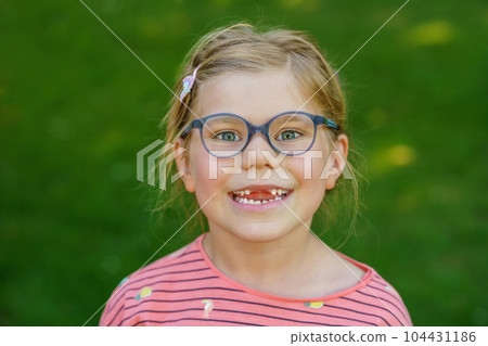 Portrait of a cute preschool girl with eye glasses and big teeth gap outdoors in park. Happy funny child on sunny summer day. Kid loosing milk tooth Portrait of a cute preschool girl with eye glasses and big teeth gap outdoors in park. Happy funny child on sunny summer day. Kid loosing milk tooth 104431186