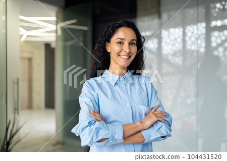 Portrait of happy and successful business woman, boss in shirt smiling and looking at camera inside office with crossed arms, Hispanic woman with curly hair in corridor. Portrait of happy and successful business woman, boss in shirt smiling and looking at camera inside office with crossed arms, Hispanic woman with curly hair in corridor. 104431520