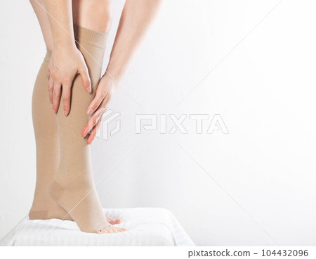 A woman puts on compression stockings for legs with varicose veins. Pain and swelling in the legs, phlebology. White background, close-up. Bloodstream A woman puts on compression stockings for legs with varicose veins. Pain and swelling in the legs, phlebology. White background, close-up. Bloodstream 104432096