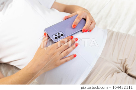 A pregnant girl holds a mobile phone near her belly. The concept of harm and deviations in the development of the unborn child. The negative impact of cellular radiation on pregnant women. 104432111