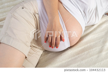 A girl in white shorts and a T-shirt lies and holds her stomach. Drawing pains and cramps in the abdomen. The concept of harbingers of childbirth. Close-up A girl in white shorts and a T-shirt lies and holds her stomach. Drawing pains and cramps in the abdomen. The concept of harbingers of childbirth. Close-up 104432113