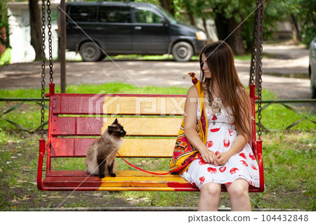 Color point cat being suspicious of its owner. Young lady trying to earn trust of her cat. An animal and a human sitting on a swing in a park, looking at each other inquiringly Color point cat being suspicious of its owner. Young lady trying to earn trust of her cat. An animal and a human sitting on a swing in a park, looking at each other inquiringly 104432848