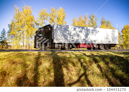 A truck with a refrigerated semi-trailer transports perishable goods against the backdrop of a forest along a country road. Copy space for text A truck with a refrigerated semi-trailer transports perishable goods against the backdrop of a forest along a country road. Copy space for text 104433270
