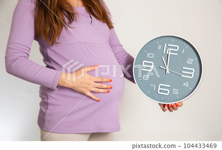 A pregnant girl in the 3rd trimester holds a wall clock against the background of a large belly. The concept of preterm birth, precursors and time to give birth. 104433469