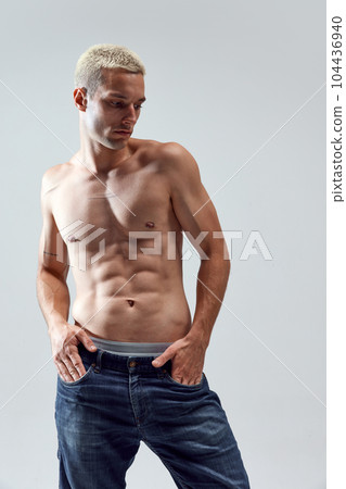Portrait of handsome young man with blonde hair, musuclar fit body posing shirtless in jeans against grey studio background 104436940