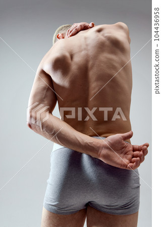 Rear view image of shirtless, muscular male body, relief back against grey studio background. Young model posing in underwear. Rear view image of shirtless, muscular male body, relief back against grey studio background. Young model posing in underwear. 104436958