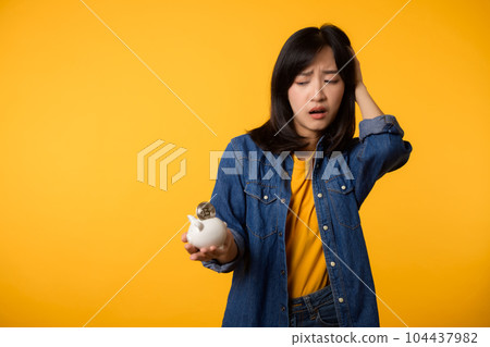 Unhappy asian young woman wearing yellow t-shirt denim shirt pulling digital coin crypto currency out of piggy bank isolated on yellow background. Payment digital money debt financial concept. Unhappy asian young woman wearing yellow t-shirt denim shirt pulling digital coin crypto currency out of piggy bank isolated on yellow background. Payment digital money debt financial concept. 104437982