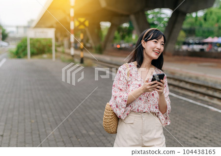 Asian young woman traveler with weaving basket using a mobile phone beside railway train station in Bangkok. Journey trip lifestyle, world travel explorer or Asia summer tourism concept. 104438165