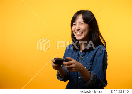 Portrait young asian woman with happy success smile wearing denim clothes holding joystick controller and playing video game. Fun and relax hobby entertainment lifestyle concept. 104438300