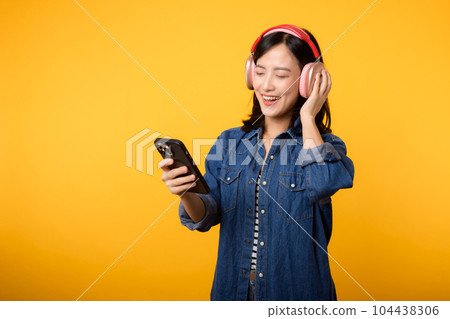 Portrait young asian cheerful woman enjoy listening audio by smartphone music application against yellow. Happy smiling female person with headphone. Sound, leisure, lifestyle, technology concept 104438306