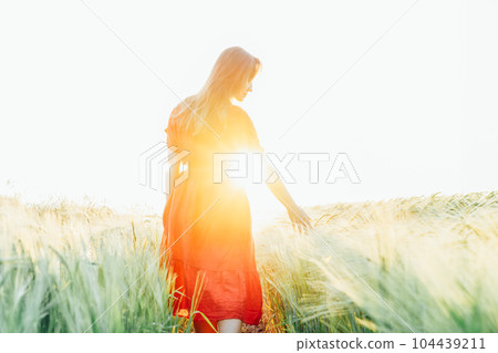 Back view woman in red dress walking and relaxing in a wheat field in sunset light with sun rays. A simple pleasure for mental health. Nature relaxation. Summertime. Soft selective focus. Copy space. 104439211