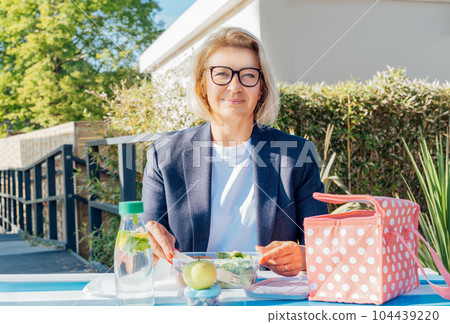 Portrait of smiling middle-aged business woman having lunch at the park outdoor cafe, picnic area during her break. Take away balanced diet lunch box with salad. Healthy eating habits and well-being 104439220