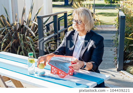 Middle-aged business woman pulls out her lunch meal at the park outdoor cafe, picnic area during her break. Takeaway balanced diet lunch box with salad. Healthy eating habits and well-being 104439221