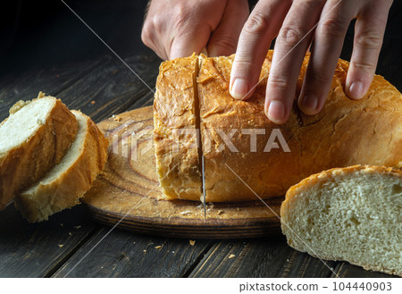 The chef cuts fresh bread with a knife on a kitchen cutting board for lunch. Healthy food and traditional bakery concept 104440903