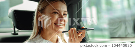 Businesswoman seated in car backseats recording voice message on phone on the way to work Businesswoman seated in car backseats recording voice message on phone on the way to work 104440950