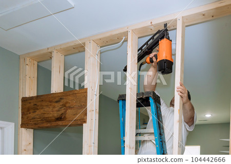 With help of an air hammer, builder nails wooden beams to frames house for construction With help of an air hammer, builder nails wooden beams to frames house for construction 104442666