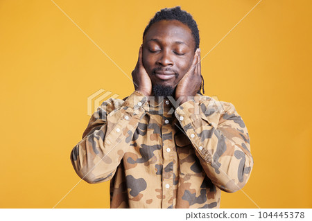 Man with close eyes covering ears with palms, doing three wise monkeys gesture in front of camera over yellow background. African american person not listening to noise and not speaking in studio 104445378