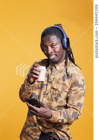 Positive smiling adult holding cup of coffee, holding smarphone scrolling to social media during lesiure time in studio. African american man searching information on internet using mobile phone Positive smiling adult holding cup of coffee, holding smarphone scrolling to social media during lesiure time in studio. African american man searching information on internet using mobile phone 104445389