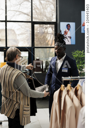 Senior customer discussing black bag material with worker, shopping for casual wear in modern boutique. Elderly client buying fashionable clothes and stylish accessories in clothing store 104445403