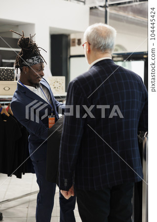 Store employee helping client choosing formal suit in showroom, african american worker helping customer to buy modern clothes. Senior man buying fashionable merchandise in modern boutique Store employee helping client choosing formal suit in showroom, african american worker helping customer to buy modern clothes. Senior man buying fashionable merchandise in modern boutique 104445424