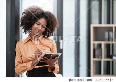 Happy African business woman chatting on digital gadgets online, holding mobile phone, using laptop computer, looking at camera, smiling. Freelance employee at home workplace. Happy African business woman chatting on digital gadgets online, holding mobile phone, using laptop computer, looking at camera, smiling. Freelance employee at home workplace. 104446618